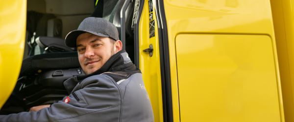 Lächelnder Lkw-Fahrer mit grauer Kappe sitzt in einem geöffneten gelben Transporter.