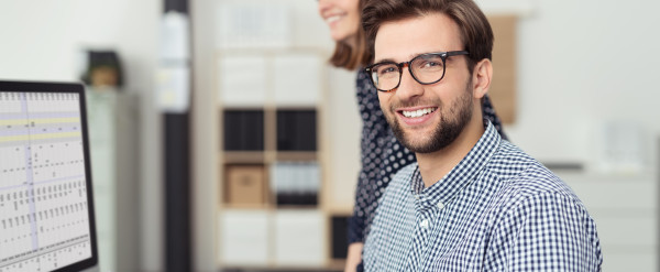 Kaufmännischer Mitarbeiter arbeitet am Computer mit Tabellenansicht, während eine Kollegin im modernen Büro unterstützt.