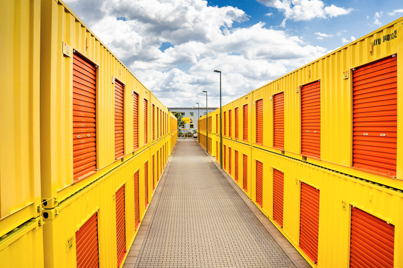 Moderne gelbe Lagercontainer mit orangenen Rolltoren in zwei Reihen, dazwischen ein gepflasterter Weg.