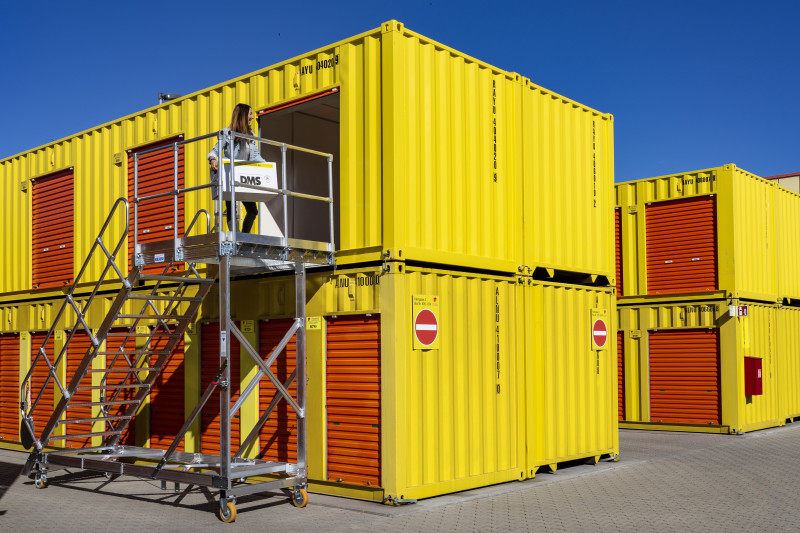 Frau auf mobiler Treppe vor gelben Lagercontainern mit orangefarbenen Rolltoren bei blauem Himmel.