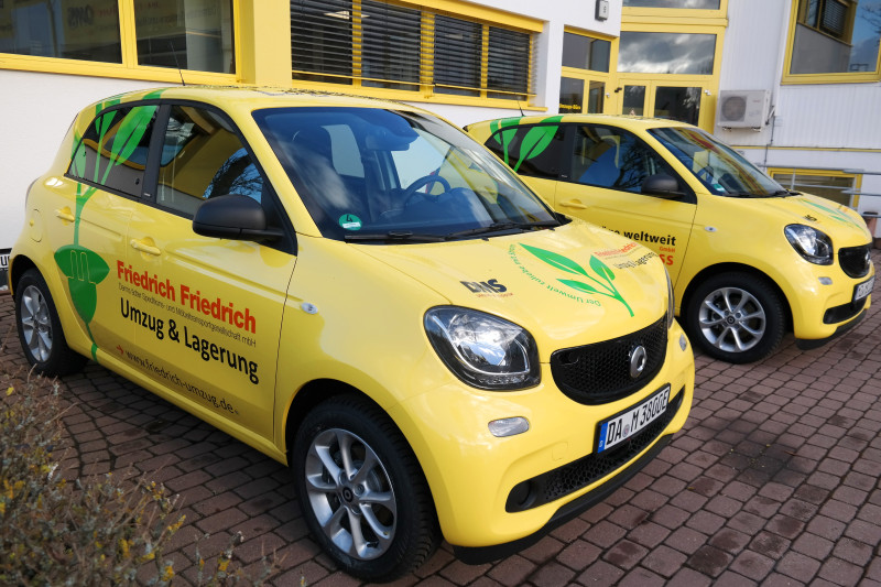 Zwei gelbe Elektroautos mit umweltfreundlicher Fahrzeugbeschriftung vor einem Firmengebäude von Friedrich Friedrich.