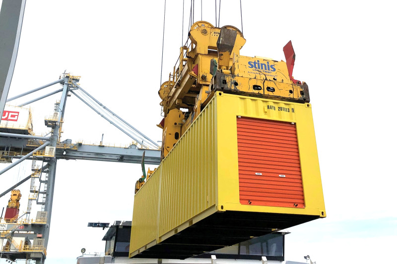 Gelber Container mit orangenen Rolltor wird mithilfe eines Krans verladen, im Hintergrund Hafenanlagen.