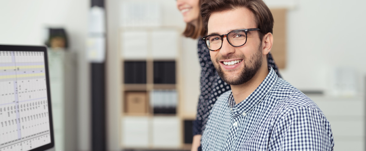 Kaufmännischer Mitarbeiter arbeitet am Computer mit Tabellenansicht, während eine Kollegin im modernen Büro unterstützt.