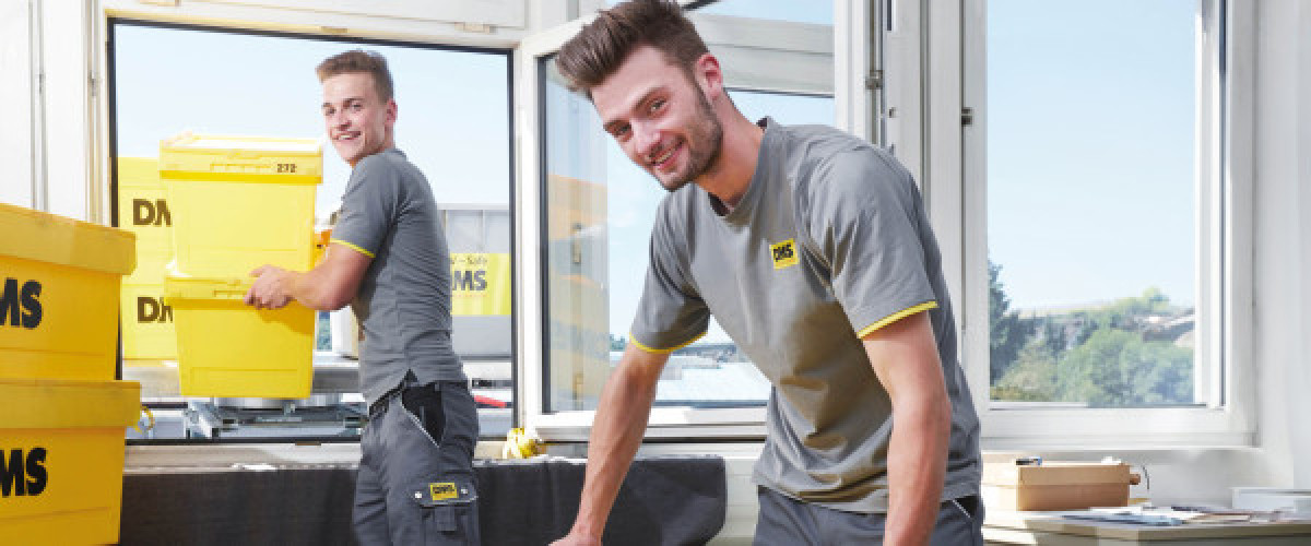 Auszubildende im Bereich Möbel-, Küchen- und Umzugsservice tragen gelbe DMS-Transportboxen und arbeiten beim Ein- und Ausladen in hellen Büroräumen.