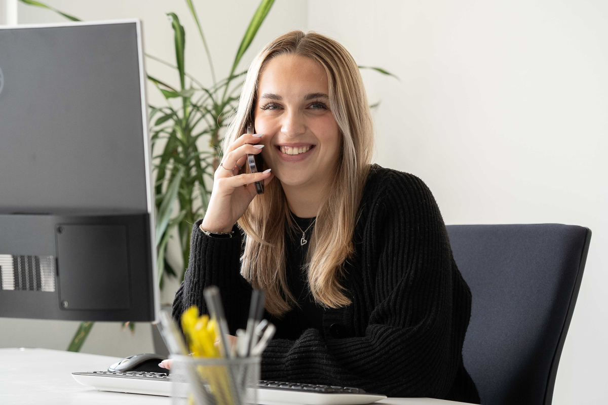 Junge Frau mit langen blonden Haaren telefoniert lächelnd am Schreibtisch im Büro.