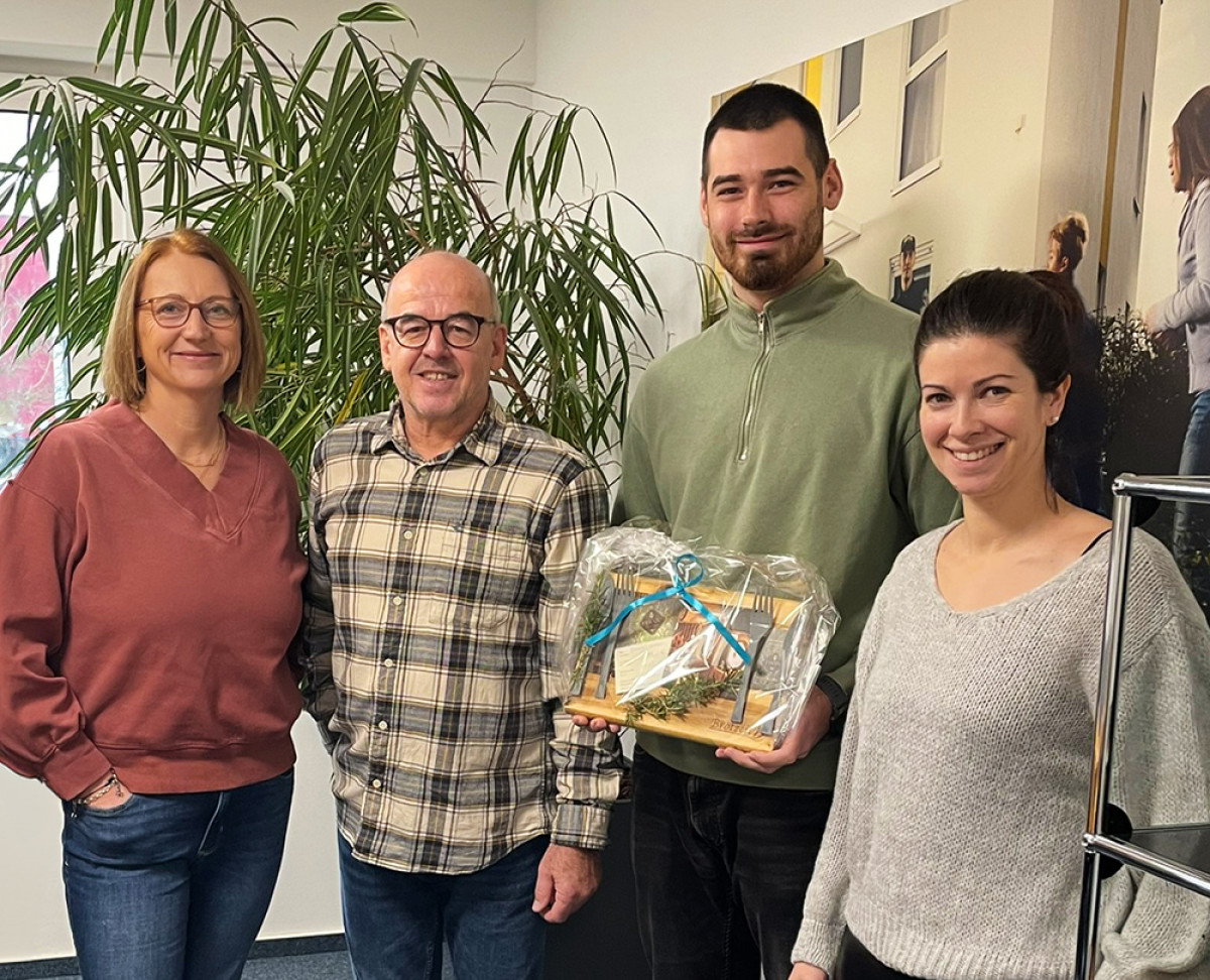 Gruppenfoto im Büro mit Geschenkübergabe, dekoriertem Präsentkorb und Pflanzen im Hintergrund.
