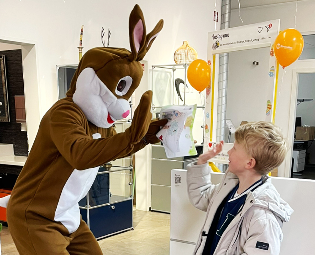 Osterhase im Kostüm gibt einem Kind ein High‑Five und begutachtet ein gemaltes Osterbild in einem Geschäft.