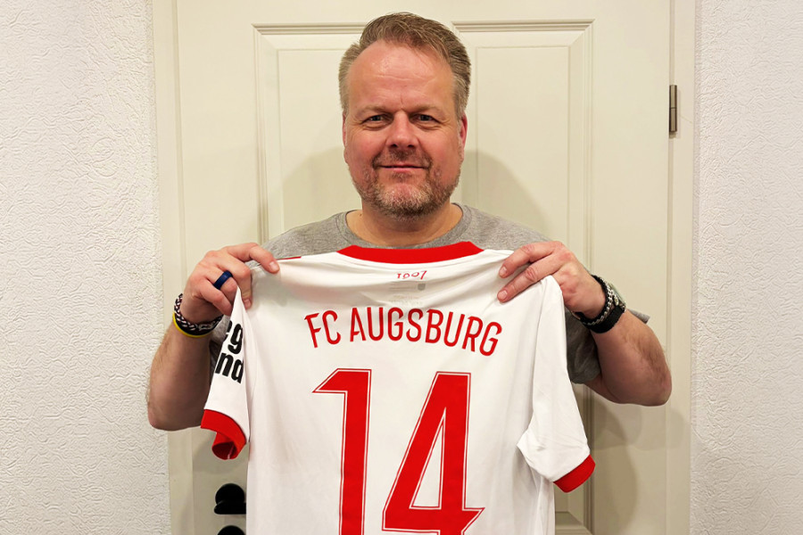 Person hält ein signiertes FC‑Augsburg‑Trikot mit der Rückennummer 14 und dem Namen Keitel vor einer Tür.