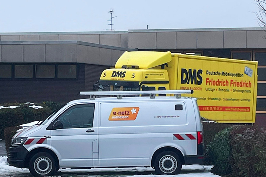 Servicefahrzeug von e‑netz Südhessen und gelber DMS‑Umzugslastwagen stehen im verschneiten Außenbereich eines Firmengebäudes.