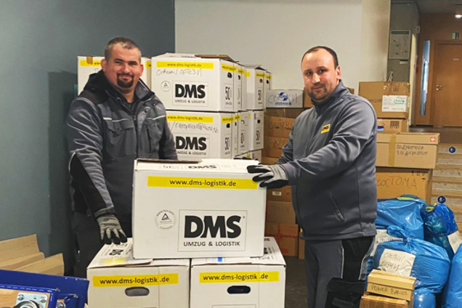 Zwei Mitarbeitende stapeln DMS‑Umzugskartons in einem Raum voller gepackter Hilfsgüter und blauer Säcke.